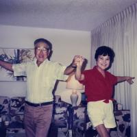 A vintage photograph of man wearing glasses dancing joyously in a living room with a woman in a red shirt. They are holding hands and their arms are up in the air.