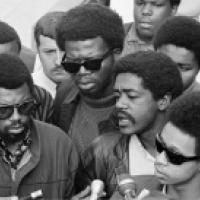A group of men stand closely together at a press conference, with microphones visible in the foreground. Their expressions and body language suggest a serious or significant public statement.