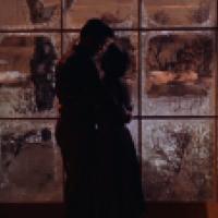 Two people stand closely together inside a building, silhouetted against a frosted window with a snowy landscape outside. Warm light filters through the panes, creating a contrast between the cold outdoors and the intimate indoor setting.