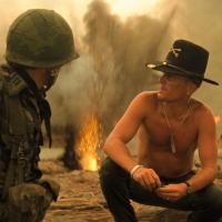 Soldiers gather in a smoky war zone with fire in the background, suggesting recent combat activity. A shirtless man in a cavalry hat speaks to another soldier, while others stand nearby in military gear.