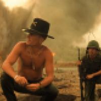 Soldiers gather in a smoky war zone with fire in the background, suggesting recent combat activity. A shirtless man in a cavalry hat speaks to another soldier, while others stand nearby in military gear.