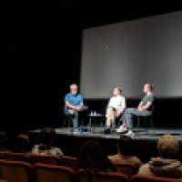 Three people sit on stage in a theater, engaged in a panel discussion or interview.