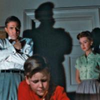 A man in a white shirt and bow tie stands with crossed arms beside a woman in a green blouse, both facing a boy in a red shirt. Dramatic shadows and tense body language suggest a moment of confrontation in a domestic setting.