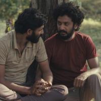 Two people sit on the ground outdoors, leaning against a tree and holding hands. The natural setting features grass and sparse vegetation, creating a quiet moment of connection.