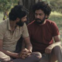 Two people sit on the ground outdoors, leaning against a tree and holding hands. The natural setting features grass and sparse vegetation, creating a quiet moment of connection.