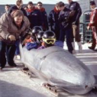 A bobsled team prepares for a run on a snowy track as a coach leans in to speak with them.