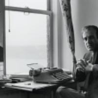  A man in a suit sits at a desk with a typewriter, papers, and books. Behind him, a window with floral curtains reveals a body of water. A mirror reflects his profile and part of the room, adding depth to the vintage office setting.