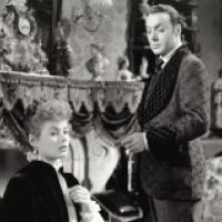 A woman in a high-collared blouse and earrings stands beside a man in a patterned velvet jacket and cravat. They are in a richly decorated Victorian room with statues, a large clock, and heavy drapery.