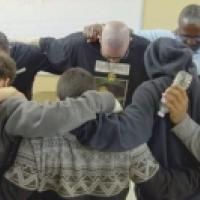 Eight people stand in a circle indoors with arms around each other’s shoulders, sharing a moment of unity. One person holds a microphone, and the background includes a whiteboard and beige walls, suggesting a classroom or community space.