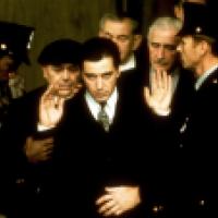 A group of people dressed in formal attire and uniforms are gathered closely together, with some raising their hands as if in greeting or acknowledgment. The scene appears to be part of an official or ceremonial event.