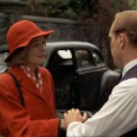 Two people are standing outdoors near a vintage car, with one person wearing a bright red coat and hat while holding hands with another person in a dark vest and white shirt.