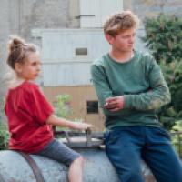 Two people sit outdoors on a large metal container in front of a rustic building. One wears a red T-shirt and gray shorts with hair in a bun; the other wears a green shirt and blue pants, holding a cigarette. Trees and bushes frame the quiet rural setting