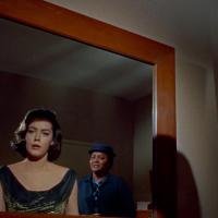 A woman in a black dress with a shimmering neckline looks into a mirror, revealing her reflection and a second person in a blue outfit and hat standing behind her. 