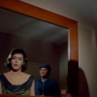 A woman in a black dress with a shimmering neckline looks into a mirror, revealing her reflection and a second person in a blue outfit and hat standing behind her. 