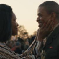 A person wearing a striped shirt gently touches the face of another person dressed in a military uniform during an outdoor gathering. People and small flags are visible in the background under a clear sky.