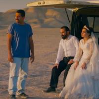 A man in a white shirt and dark pants sits beside a woman in a white wedding dress at the open back of a silver van. Another person stands nearby in a blue shirt and light pants. The desert-like landscape and mountains in the background contrast with the formal wedding attire.