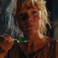 A young person with light hair holds a spoon with green gelatin, gazing intently off-camera. Dim lighting and blurred background elements, including a yellow flower and greenery, create a dramatic and contemplative atmosphere.