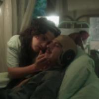 A person lies in a hospital bed being attended to by another individual; the dimly lit medical facility has green metal bed frames and curtains