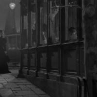 A woman in a long coat and hat walks along a cobblestone sidewalk at night past a vintage storefront with ornate windows. Inside, people are visible through the glass, and a sign partially reads “Billiard.” A streetlamp casts light on the scene, with a vi