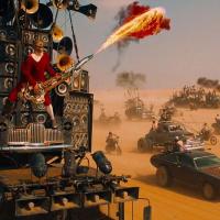A massive vehicle outfitted with speakers and horns barrels through a desert as a performer in red plays a flame-throwing guitar strapped to its front. Behind it, a convoy of weaponized cars and motorcycles kicks up dust under a blazing sky, evoking a cha