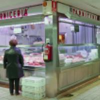 A person in a dark coat stands at the counter of a butcher shop labeled “Carnicería.” Various meats and sausages are displayed, with stacks of eggs to the right. The shop is numbered 16 and features tiled floors, fluorescent lighting, and a fire extinguis