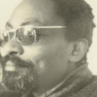 A sepia-toned portrait of a person with short curly hair, glasses, and a beard, resting their head on their hand. They wear a textured sweater and gaze thoughtfully to the side, evoking a quiet, introspective mood.