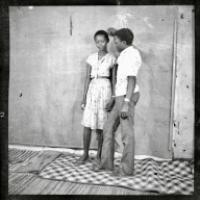 A black-and-white photograph shows two people indoors against a textured backdrop. One stands barefoot on a woven mat wearing a patterned dress, while the other leans slightly toward them with one arm raised, dressed in a short-sleeved shirt and trousers.