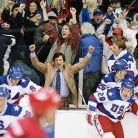Players in white USA hockey jerseys cheer from the bench as fans erupt behind them. A man in a tan jacket and tie raises his fists in celebration. The scene captures a moment of triumph and national pride during an intense game.
