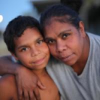 Two people embrace closely with their faces touching, looking directly at the camera. The softly blurred background suggests an outdoor setting during twilight, highlighting a moment of intimacy and connection.