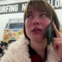 A person in a fluffy white coat holds a smartphone to their ear, appearing serious. Behind them, surfboards lean against a white brick wall and a colorful mural of a van with tropical flowers reads “SURFING NOT ALONE.” 
