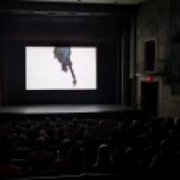 Inside a darkened movie theater, audience members watch a film showing an upside-down image of the Statue of Liberty’s torch and crown.