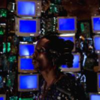 A person stands in front of a wall of old CRT monitors, each displaying a solid blue screen. They wear elaborate accessories made from electronic components, with coils in their hair and circuit boards on their clothing. The background is filled with wire