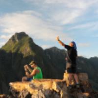 A group of people stands on a rocky mountain summit, taking photos and selfies. Behind them, lush green peaks stretch under a partly cloudy sky.