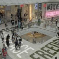 A busy shopping mall features retail stores like H&M and John Lewis, with crowds walking and dining. Bold neon slogans—“WAR IS PEACE,” “FREEDOM IS SLAVERY,” and “IGNORANCE IS STRENGTH”—are prominently displayed, evoking Orwellian themes amid the consumeri