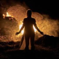 A person stands with arms slightly outstretched in front of a large fire burning hay bales at night. The flames and smoke illuminate the dark surroundings, creating a dramatic silhouette effect.