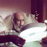 A person with long white hair sits in bed holding a document under a magnifying lamp