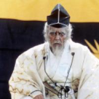An elderly man with a white beard and intense expression wears a white robe with gold patterns and a black hat trimmed in white. He stands before a large banner featuring a yellow and black sunburst design, evoking a theatrical or historical Japanese sett