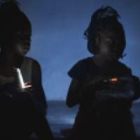 Two children sit in a dimly lit space. One holds a small light source that illuminates their face, while the other holds a portable radio with a digital display. The background is dark blue, suggesting a nighttime or off-grid setting.