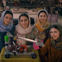 Five young people stand indoors behind a table displaying a robotic arm mounted on a wheeled base. Four women and one man wear traditional attire with headscarves and warm clothing. The background includes workshop items like an anvil, tires, and mechanic