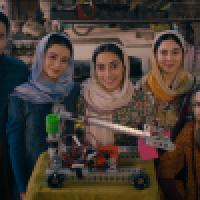 Five young people stand indoors behind a table displaying a robotic arm mounted on a wheeled base. Four women and one man wear traditional attire with headscarves and warm clothing. The background includes workshop items like an anvil, tires, and mechanic