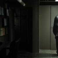 A dimly lit room shows a figure wrapped in dark, shiny material standing near a wall, while a computer monitor on a desk displays an image of a patterned garment. Clipboards are mounted on the wall to the left, and the setting appears industrial or office