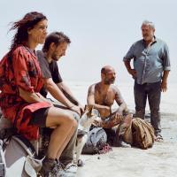 Four people and a dog gather in a desert landscape. Three sit on the ground or on bags—one in a red and black patterned outfit, one in a black shirt, and one shirtless with tattoos and colorful pants. A fourth person stands nearby in a blue shirt and jeans. Backpacks and gear surround them, and the clear sky emphasizes the stark, sandy terrain.