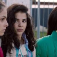 Three people stand outdoors near a metal fence, dressed in jackets and sweaters. One wears a red jacket, another has curly hair and a light hoodie, and the third wears a green jacket over a yellow turtleneck. Their serious expressions suggest a moment of 