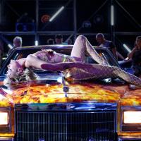 A female figure lays across the hood of a Cadillac which has flames painted on the hood. Several onlookers appear in the background.