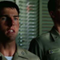 Two individuals wearing military uniforms stand indoors in front of horizontal window blinds. One uniform displays a name tag, insignia, and colorful ribbons.