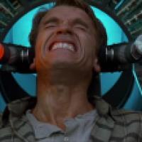 A person is seated in a futuristic chair with two large mechanical devices positioned on either side of the head, inside a high-tech environment with illuminated panels and machinery.