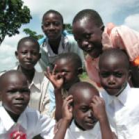 A group of children and one adult are gathered outdoors, wearing light-colored clothing, with trees and a blue sky in the background. They appear to be posing closely together for a photo.
