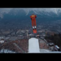 A ski jumper in a blue suit is airborne above a snowy ramp, with a large crowd in a stadium below and a cityscape surrounded by mountains in the background. The scene captures the moment of flight during a ski jumping event.