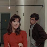 Three people stand in a hallway with white walls and a green door, engaged in a tense conversation. The central figure in red gestures while the others listen.