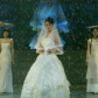 Three people wearing elegant white dresses stand on a stage with falling snow, while two hold parasols. The central figure is dressed in a detailed bridal gown, creating a dramatic winter-themed scene.